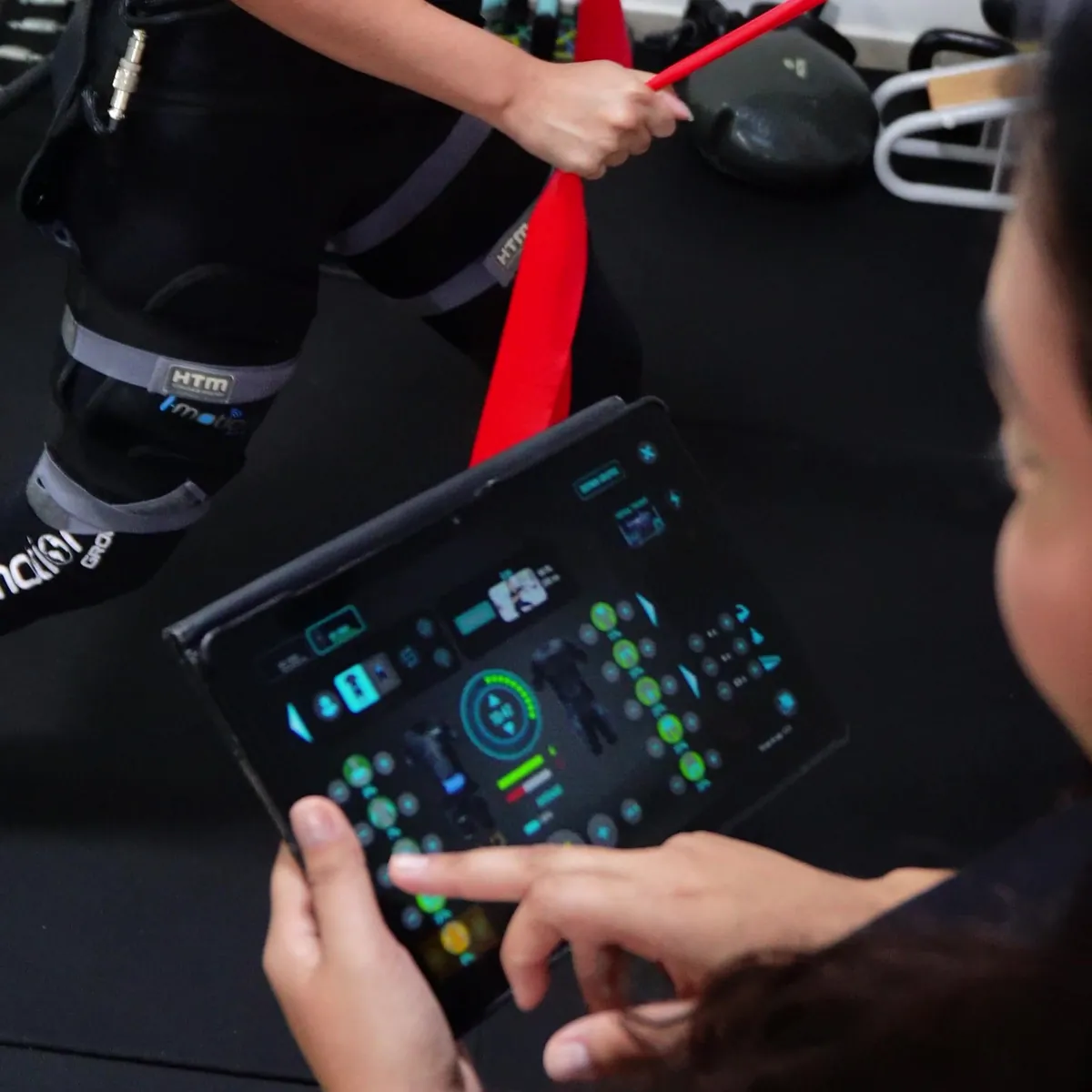 Profissional ajustando no tablet a intensidade da eletroestimulação enquanto a aluna realiza um exercício